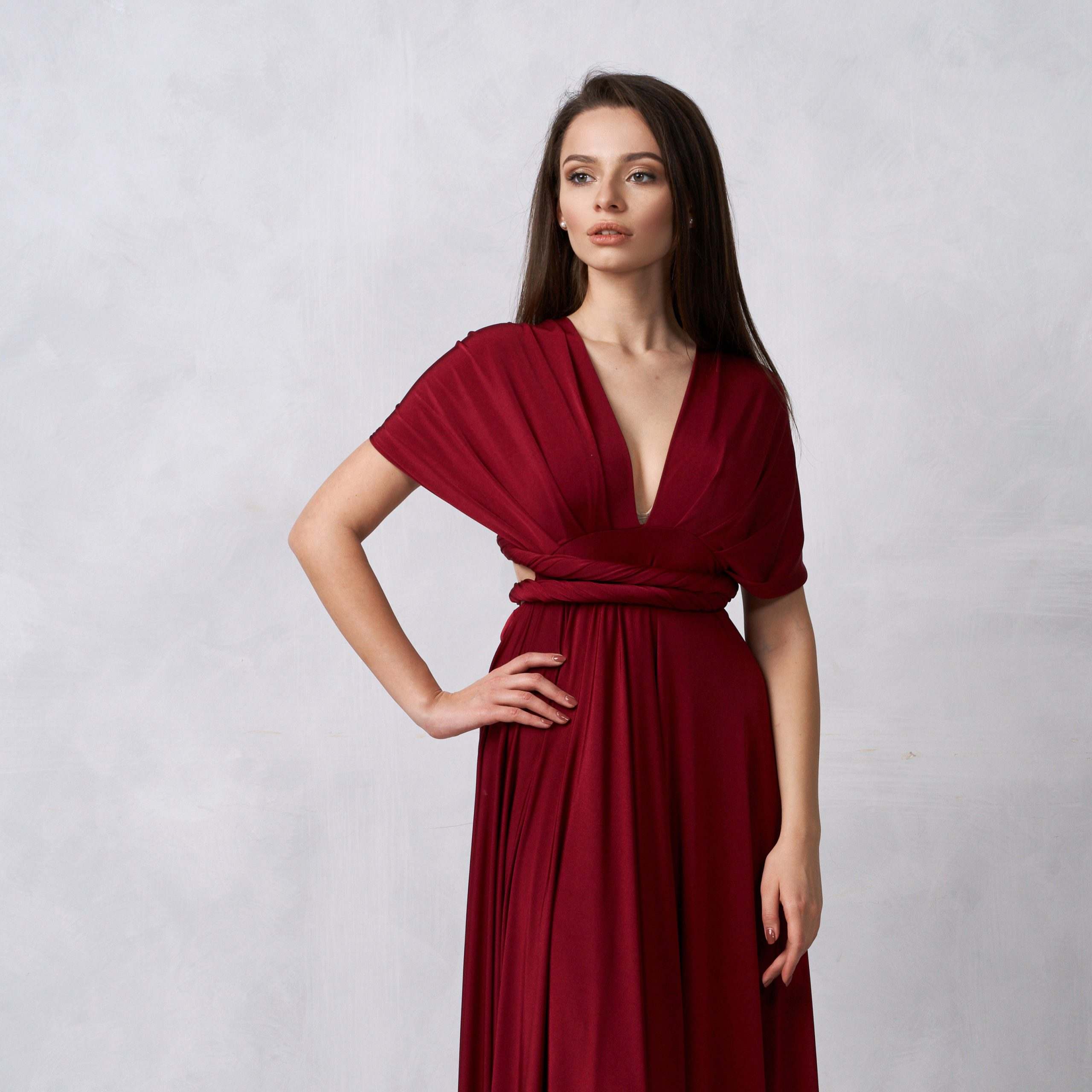 Beautiful long haired young woman dressed in stylish cherry red bandeau maxi dress posing against white wall on background. Elegant brunette female model demonstrating evening outfit in studio.